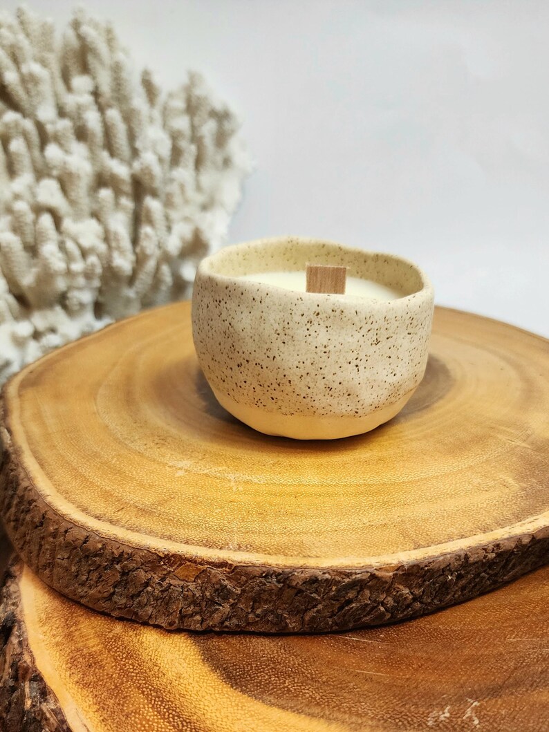 Op de afbeelding: Een gespikkelde witte keramische kaars pot met een houten lont, die op een houten schijf staat.