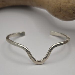 May include: A silver-toned cuff bracelet featuring a distinct V-shaped center dip and a textured, hammered finish. The open-ended design has smooth, rounded tips and a reflective metallic surface that catches the light across its curved form.