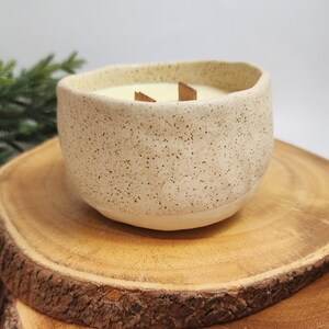 Op de afbeelding: Een witte keramische kaars pot met een gespikkelde afwerking. De kaars is aangestoken en heeft een houten lont.