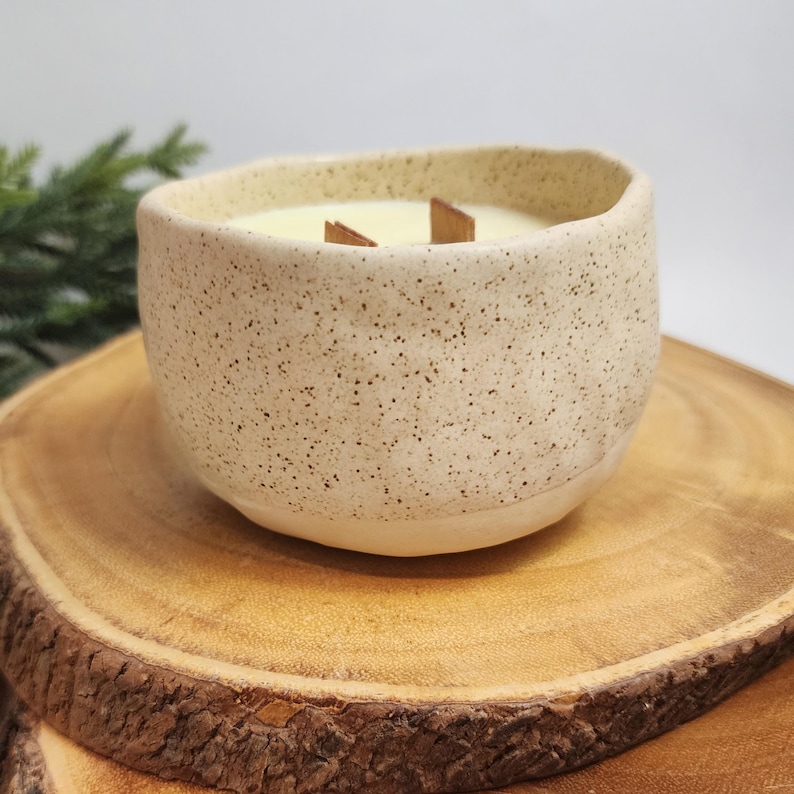 Op de afbeelding: Een witte keramische kaars pot met een gespikkelde afwerking. De kaars is aangestoken en heeft een houten lont.