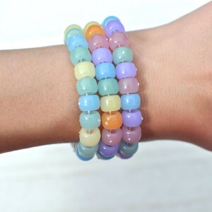 May include: Three pastel-colored beaded bracelets. The bracelets are made of round beads in shades of blue, green, yellow, orange, and purple. The bracelets are stacked on top of each other.