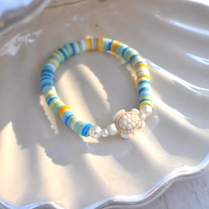 May include: A colorful bracelet featuring a turtle charm. The bracelet is made of blue, yellow, and white beads. The turtle charm is off-white and has a detailed shell design. The bracelet is accented with small, white pearl beads.