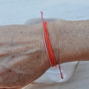 May include: A handmade friendship bracelet featuring strands of pink, orange, and red thread. The bracelet is tied around a wrist, showcasing the vibrant colors and simple design. This accessory is perfect for everyday wear.