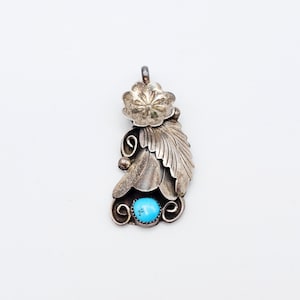 Puede incluir: Colgante de plata con diseño floral y de hojas, con una piedra turquesa brillante. El colgante tiene una flor en la parte superior, una hoja detallada y remolinos decorativos. La piedra turquesa está engastada en un bisel de plata.
