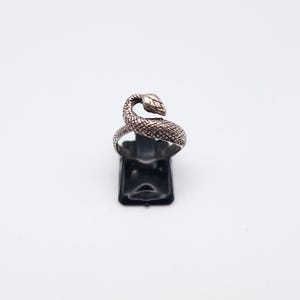 May include: A silver-coloured ring shaped like a snake. The snake's head is raised, and its body coils around the finger. The ring is displayed on a black stand against a white background. The ring's design features textured scales.