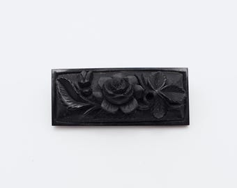 Peut inclure: Un élément décoratif rectangulaire noir avec un motif floral sculpté. Le motif comprend une rose centrale, des feuilles et d'autres éléments floraux. La pièce est probablement faite d'un matériau sombre et solide.