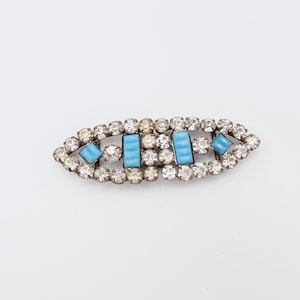 May include: An oval-shaped brooch with a silver-tone metal setting. The brooch features clear, round rhinestones around the perimeter and light blue rectangular and square stones in the center. A vintage-style accessory.