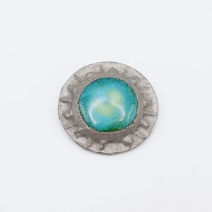 May include: A circular brooch featuring a central, smooth, domed cabochon with a gradient of turquoise, blue, and yellow-green hues. The center is encased in a textured, hammered silver-toned metal frame with a scalloped, irregular edge.