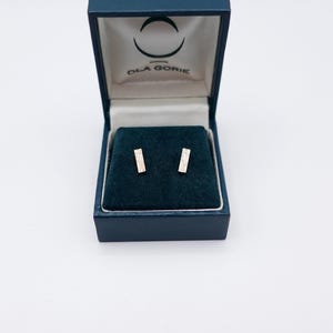 May include: A navy blue jewellery box, open to reveal a pair of small, rectangular stud earrings. The earrings are displayed on a dark green velvet-like surface. The box is branded with the name "OLA GORIE" on the inside of the lid.