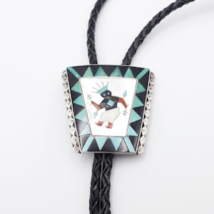 May include: A silver bolo tie with a black leather cord. The rectangular pendant features a central image of a Native American figure in a colorful mosaic design, framed by black, turquoise, and silver accents. The bolo tie is a Southwestern style accessory.