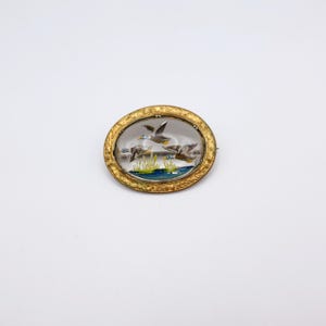 Puede incluir: Broche ovalado dorado con un centro transparente y abovedado que representa tres patos volando sobre un estanque con juncos. El broche tiene un borde ornamentado y una cierre de alfiler. La escena está pintada en el interior.