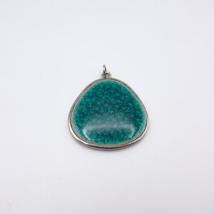 May include: A teal-colored pendant with a silver-colored border. The pendant has a triangular shape with a textured surface. The bail is silver-colored. The pendant is likely a piece of jewelry.