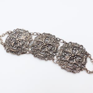 May include: Silver metal belt featuring three decorative, floral-patterned rectangular panels linked by a chain. The belt has a vintage appearance, with intricate filigree and a tarnished finish, ideal for adding a touch of elegance.