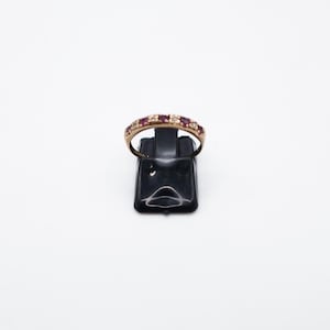 Könnte beinhalten: Ein goldfarbener Ring mit einem Band aus abwechselnd roten und klaren Steinen. Der Ring wird auf einem schwarzen Schmuckständer vor weißem Hintergrund präsentiert. Die Steine sind gleichmäßig angeordnet.