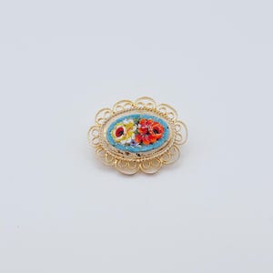 Peut inclure: Broche ovale dorée avec un motif floral en mosaïque. La mosaïque présente des fleurs rouges, jaunes et blanches sur un fond bleu clair, encadrées par une délicate bordure en filigrane doré. Un accessoire vintage.
