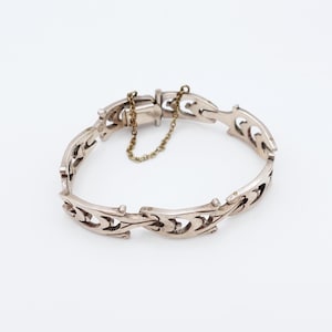 May include: Silver-toned bracelet with an ornate, openwork design. The bracelet features a series of interconnected links, each with a repeating, stylized pattern. A small chain secures the clasp. The jewelry is displayed against a white background.