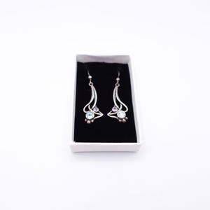 May include: A pair of silver drop earrings featuring a curved, openwork design with a small round light blue gemstone at the base and a tiny clear stone accent above, presented in a white rectangular box with a black velvet insert.