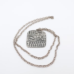May include: A silver-toned pendant necklace with a square pendant featuring etched figures and symbols. The chain is a matching silver color. The pendant has a textured surface with various etched designs, including human-like figures, animals, and geometric shapes.