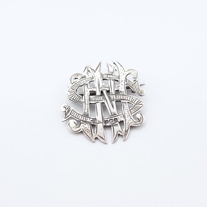 May include: A silver brooch with an intertwined monogram design. The brooch has an ornate, openwork pattern with the letters "N" and "S" visible. The metal has a polished finish, and the brooch is set against a white background.