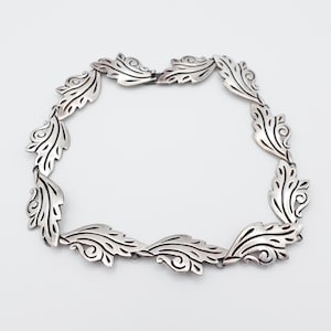 May include: A silver-toned necklace featuring a repeating leaf design. Each leaf has detailed veining and a curled accent. The necklace is connected by small links, creating a flexible and elegant piece of jewelry.