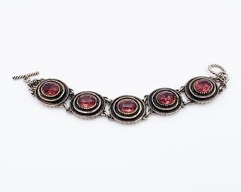 May include: Silver-toned bracelet featuring five circular settings, each holding a round, reddish-pink gemstone. The bracelet has a toggle clasp closure. The settings are detailed with a textured border, and the links are delicate.