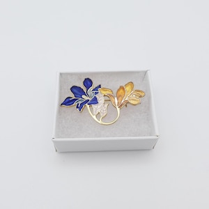 Puede incluir: Broche floral con flores de esmalte azul y amarillo y detalles en metal dorado. El broche se presenta en una caja de regalo blanca con interior texturizado, ideal para regalar.