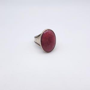 May include: A silver ring with a large, oval, deep pink gemstone. The ring's band has a textured, rope-like design on the sides. The gemstone is smooth and polished, set in a simple bezel.