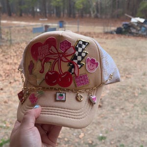May include: Tan trucker hat with a mesh back and a curved brim. The hat is decorated with patches including cherries, a lightning bolt, and a pink cowboy hat. The text "Bless Your Heart" is visible on the front.