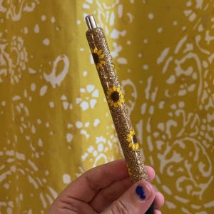 Puede incluir: Un bol&iacute;grafo cil&iacute;ndrico con acabado de purpurina dorada decorado con un patr&oacute;n de peque&ntilde;os girasoles amarillos. El bol&iacute;grafo tiene una punta de metal plateado en la parte superior y una punta de tinta negra en la parte inferior con textura brillante.
