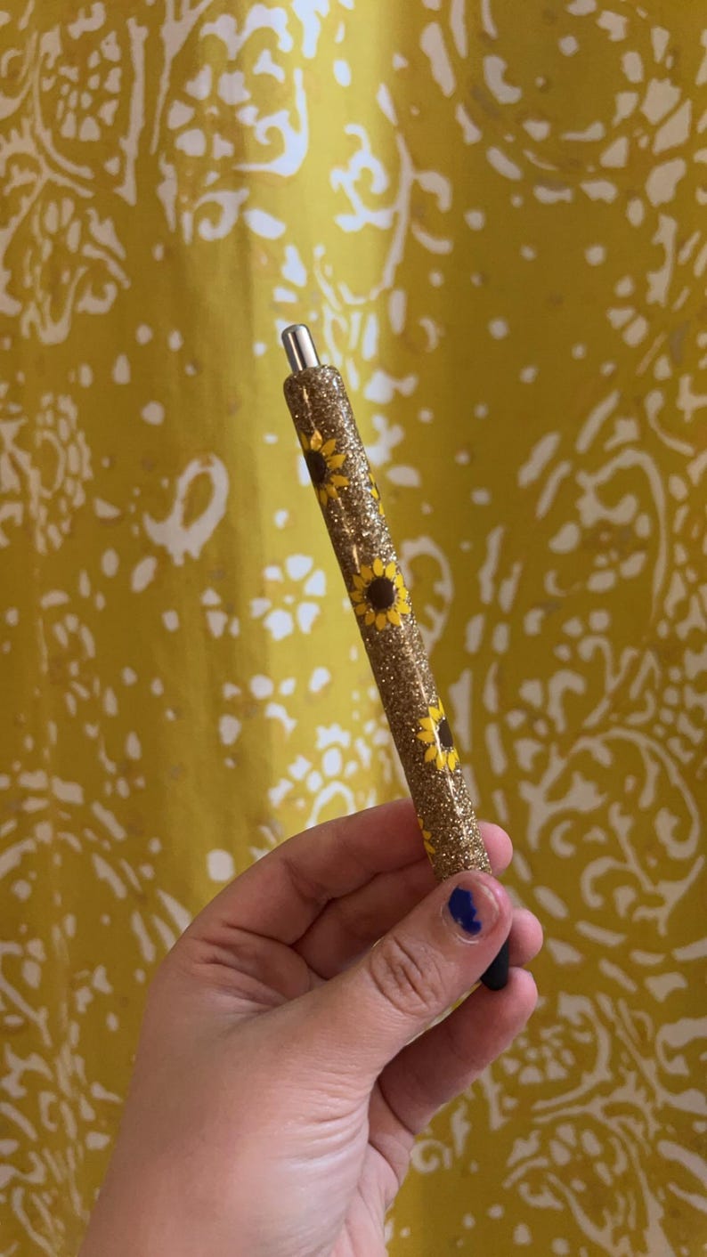 Puede incluir: Un bol&iacute;grafo cil&iacute;ndrico con acabado de purpurina dorada decorado con un patr&oacute;n de peque&ntilde;os girasoles amarillos. El bol&iacute;grafo tiene una punta de metal plateado en la parte superior y una punta de tinta negra en la parte inferior con textura brillante.
