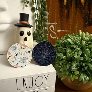 May include: Halloween-themed decorations featuring a ghost figure with a top hat and two round coasters. One coaster has a spiderweb design, and the other has a ghost and spiderweb pattern. The decorations are on a white surface.