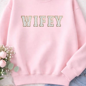 May include: A light pink sweatshirt with the word "WIFEY" in gold and white block letters. The sweatshirt has a crew neck and long sleeves. The image also shows a pair of blue denim shorts and a bouquet of flowers.