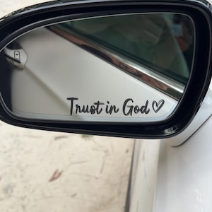 May include: A white car side mirror with a black frame. The mirror displays the text "Trust in God" in a cursive font, followed by a small heart symbol. The car is parked outdoors, with a blurred background of trees and a sandy surface.