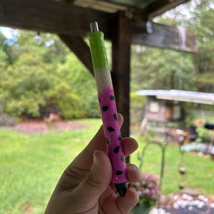 May include: A pink and green glitter pen with black watermelon seeds. The pen is held in a hand.