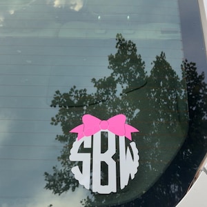May include: A white car window decal featuring a pink bow above a white monogram with the letters SBW. The decal is circular with a scalloped edge. The car window reflects a cloudy sky and trees.
