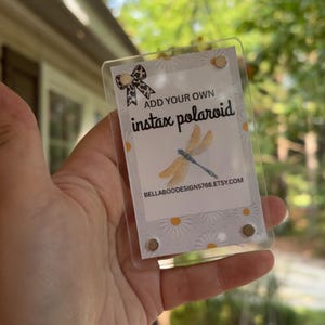 May include: Clear plastic Instax Polaroid frame with a printed insert. The insert features a dragonfly illustration, daisy pattern, and text that reads "ADD YOUR OWN instax polaroid" and "BELLABOODESIGNS768.ETSY.COM".