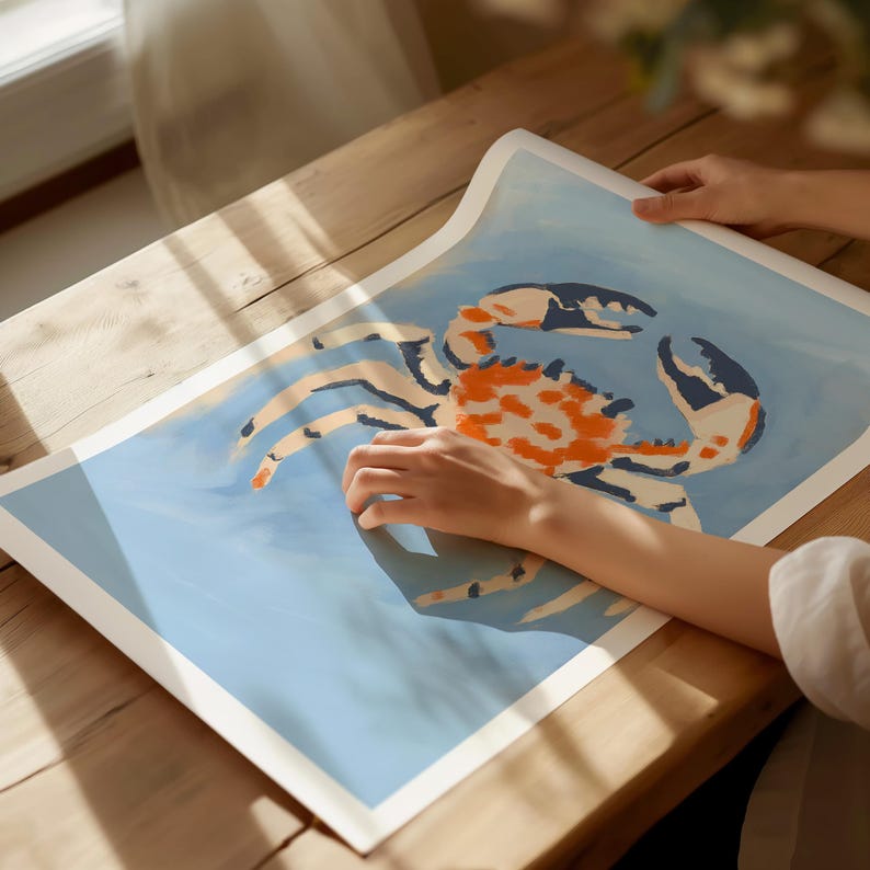 May include: A blue and white print of a crab with orange and white markings. The crab is painted in a stylized, almost cartoon-like way. The print is on a white background and is being held by a hand.