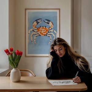 May include: A framed print of a stylized orange and white crab on a blue background. A person is sitting at a table with a vase of red tulips and is drawing on a piece of paper.