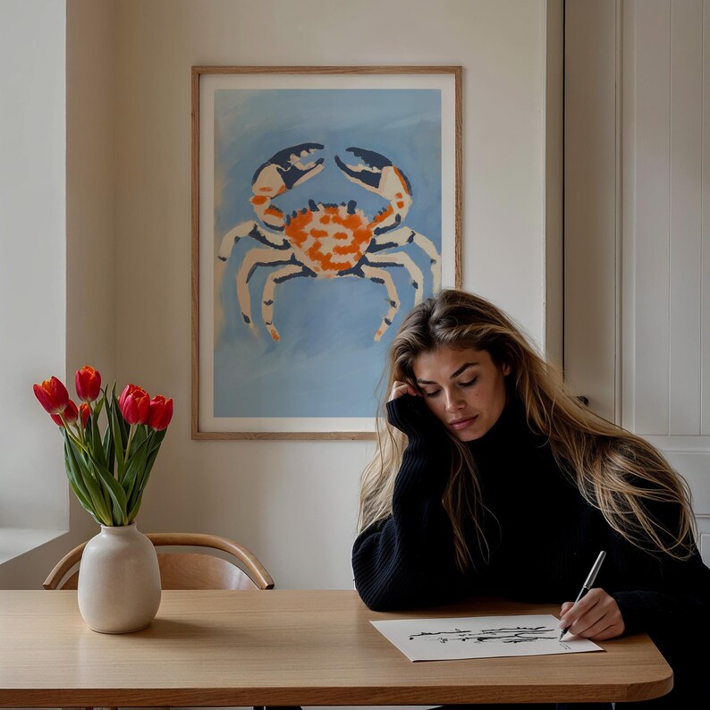 May include: A framed print of a stylized orange and white crab on a blue background. A person is sitting at a table with a vase of red tulips and is drawing on a piece of paper.