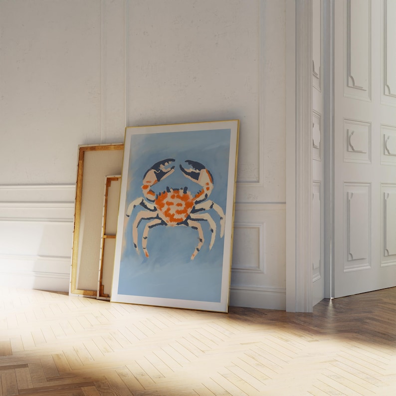 May include: A framed print of a colorful crab on a light blue background. The crab is orange, white, and black. The frame is gold.