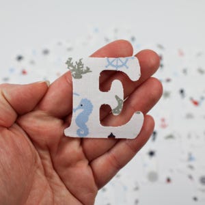 May include: A white fabric letter E with a blue and gray nautical pattern. The letter is cut out and held in a hand.