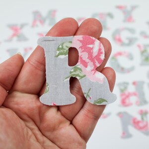 Op de afbeelding: Een grijze stoffen letter R met een bloemenpatroon in roze en groen. De letter wordt in een hand gehouden.