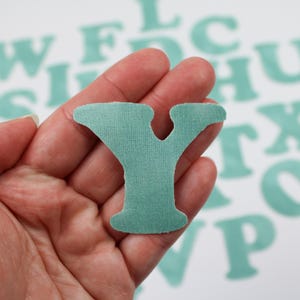 Op de afbeelding: Een turquoise viltletter Y, vastgehouden in een hand, met andere turquoise viltletters op de achtergrond.