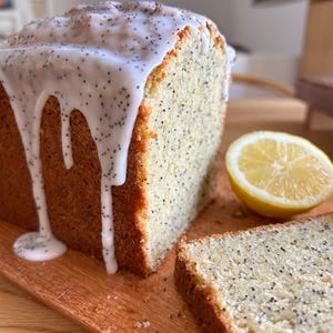 May include: A loaf of lemon poppy seed cake with white icing drizzled over the top. A slice of the cake is cut and placed next to the loaf. A lemon half is also on the wooden cutting board.