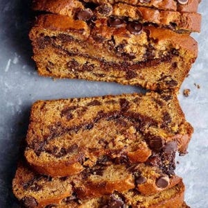 May include: Sliced loaf of chocolate chip bread. The bread has a golden-brown crust and is filled with swirls of chocolate. Chocolate chips are scattered on top of the bread. The bread is stacked on a light gray surface.