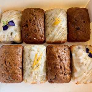 May include: A white cardboard box containing eight rectangular mini loaves of bread. The assortment includes brown loaves with visible chocolate chips, light-coloured loaves with poppy seeds and white glaze, and loaves topped with edible purple flowers.