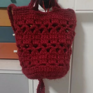 Könnte beinhalten: Eine kleine, gehäkelte Tasche in burgunderrot. Die Tasche hat einen langen Riemen und hängt an einem schwarzen Haken.