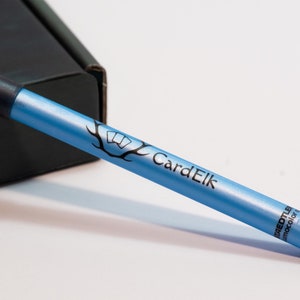 Può includere: Uno strumento blu per mescolare le carte con il testo "CardElk" e "PREDTUBE" stampato su di esso. Lo strumento ha una punta in gomma nera e un'impugnatura nera.