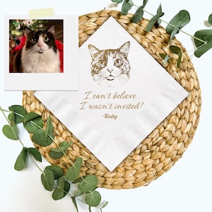 May include: White square napkin with a gold cat illustration and the text "I can't believe I wasn't invited! -Ruby". A polaroid photo of a cat is in the upper left corner. The napkin is on a woven placemat.