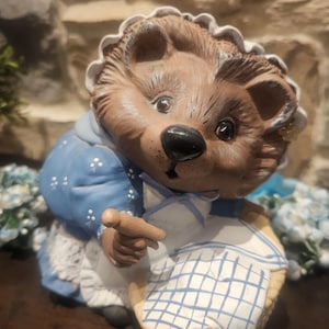 May include: A ceramic hedgehog figurine wearing a blue dress with white polka dots and a white apron with a blue checkered pattern. The hedgehog has a brown face and is wearing a white bonnet.